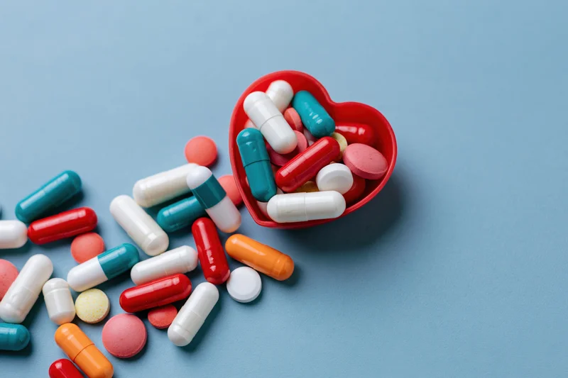 Assorted pharmaceutical capsules and tablets spilling from a red heart-shaped container on a blue surface