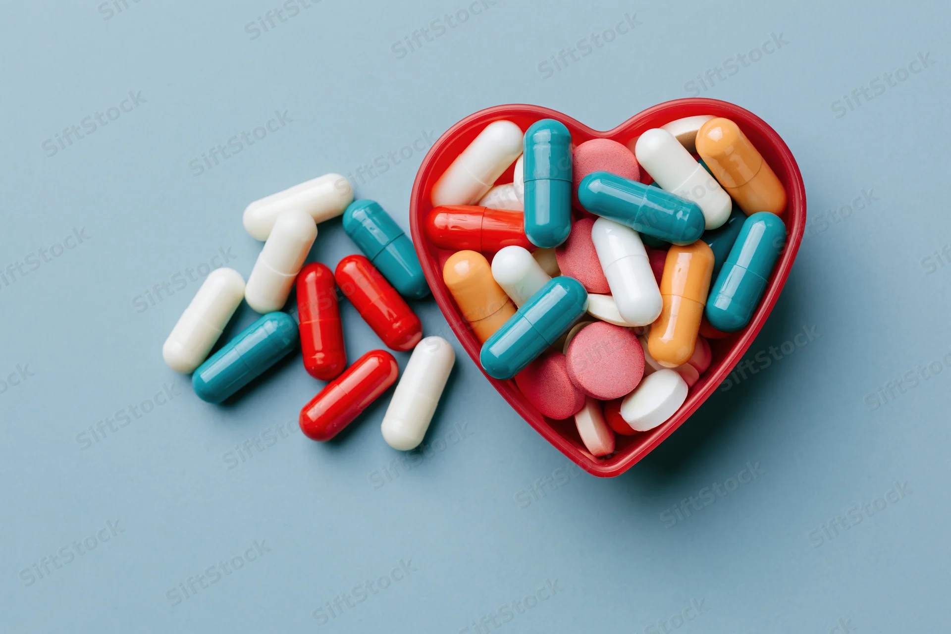 Assorted pharmaceutical capsules and tablets spilling from a red heart-shaped container on a blue surface