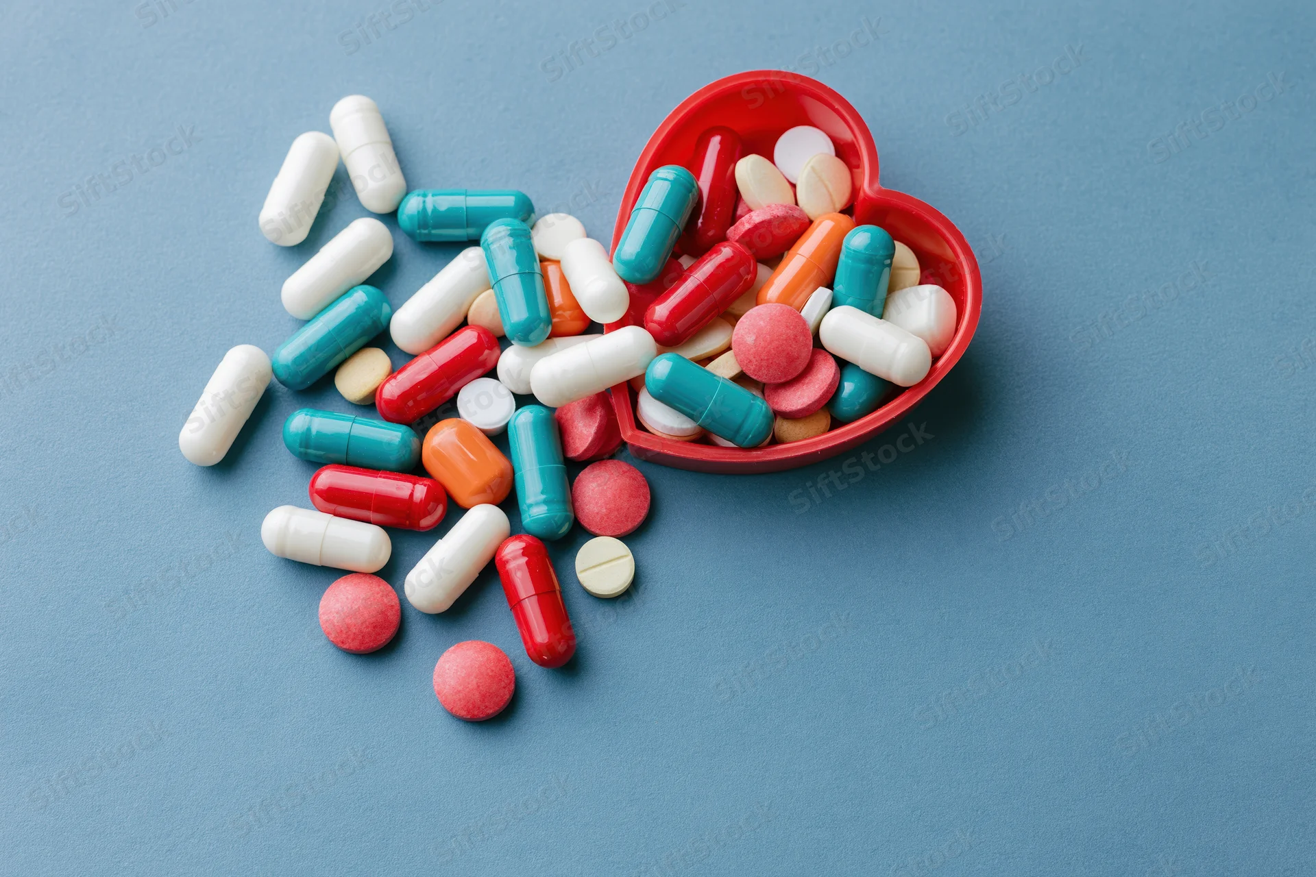 Assorted pharmaceutical capsules and tablets spilling from a red heart-shaped container on a blue surface