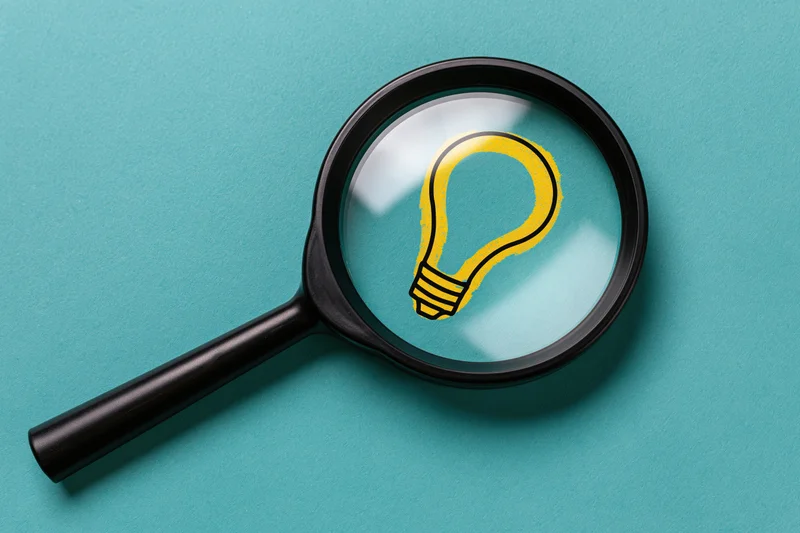 Black magnifying glass with yellow lightbulb symbol on teal paper background