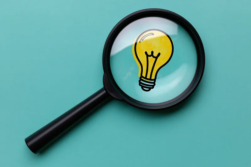 Black magnifying glass resting on teal surface with illustrated lightbulb visible through lens