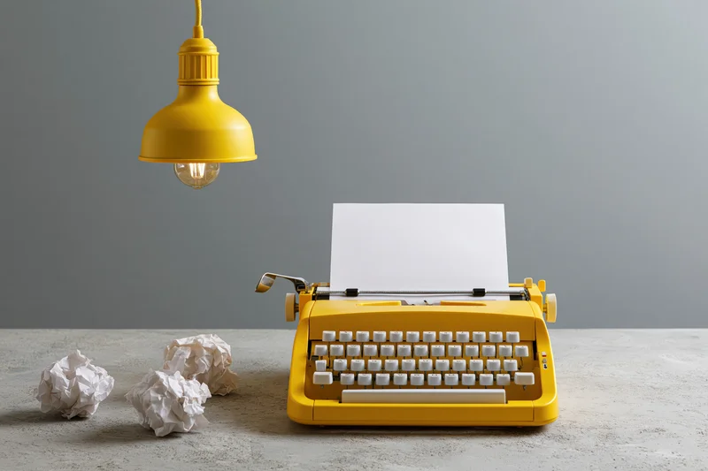 Yellow vintage typewriter with blank paper on desk under hanging lamp, crumpled papers beside it
