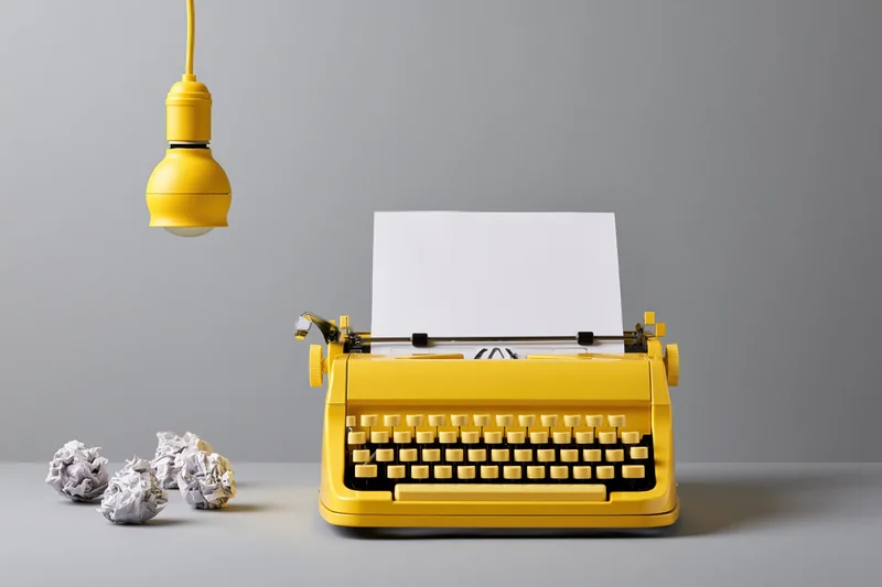 Yellow vintage typewriter with blank paper and crumpled drafts on a desk under a hanging lamp