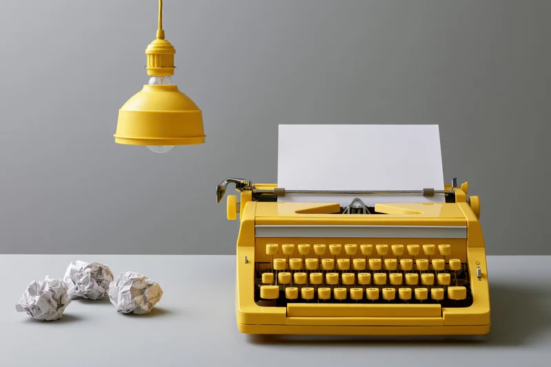 Yellow vintage typewriter with blank paper and crumpled drafts on a desk under a hanging lamp