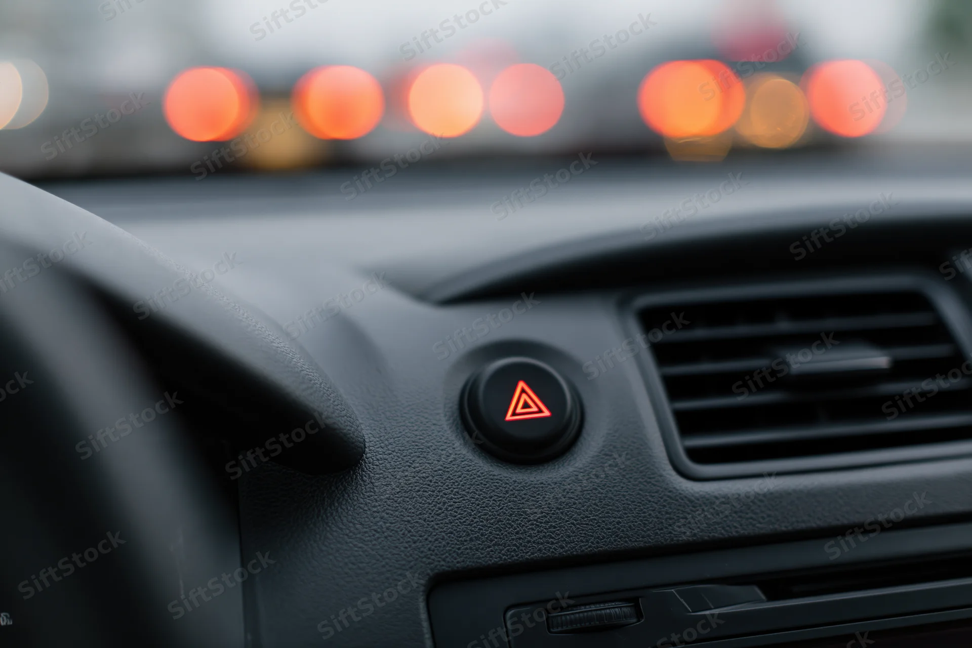 Close-up of car dashboard console showing illuminated hazard light button and air vent with blurred traffic lights in background