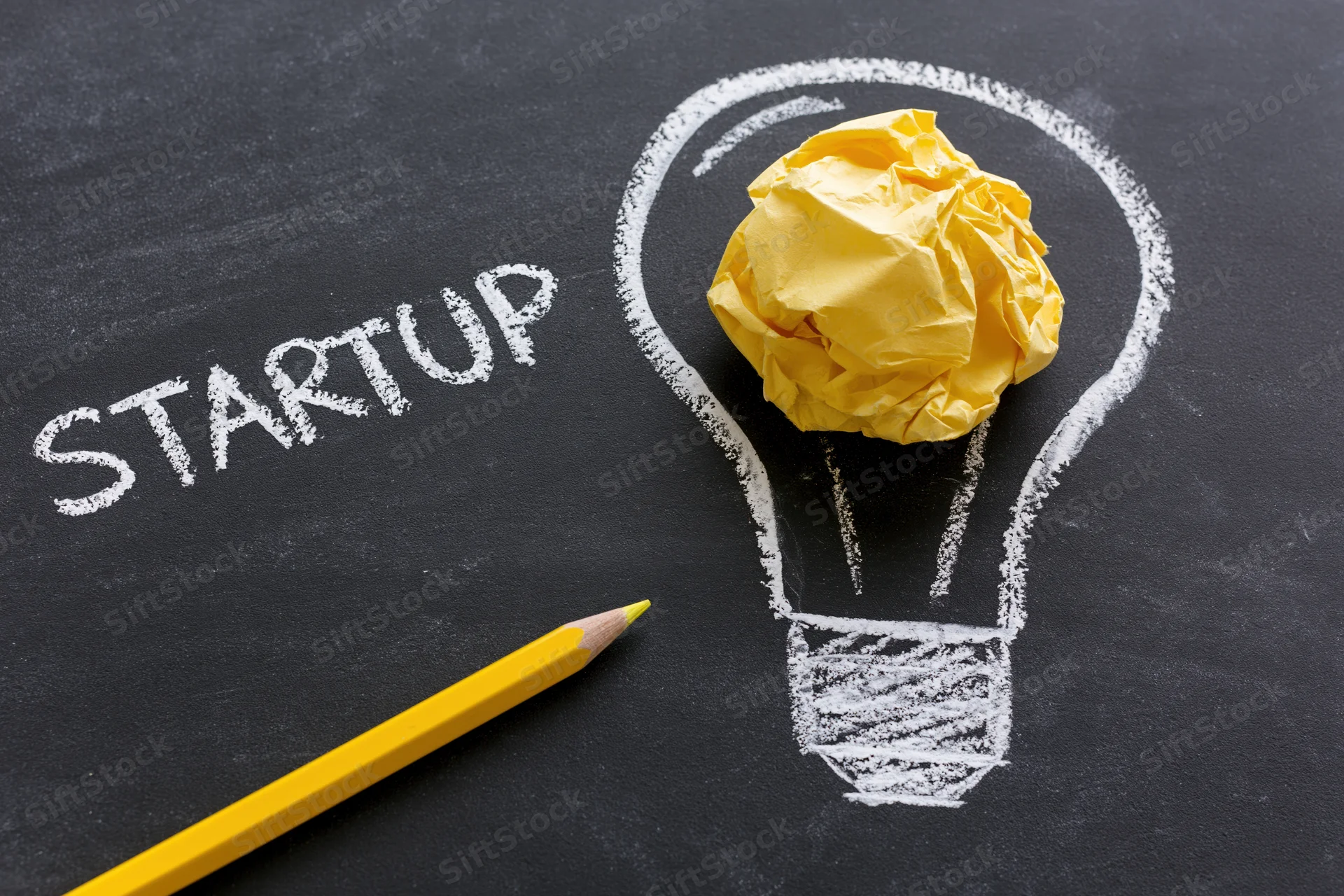 Chalk-drawn lightbulb with crumpled yellow paper inside and the word startup written beside it on a blackboard, with a yellow pencil nearby