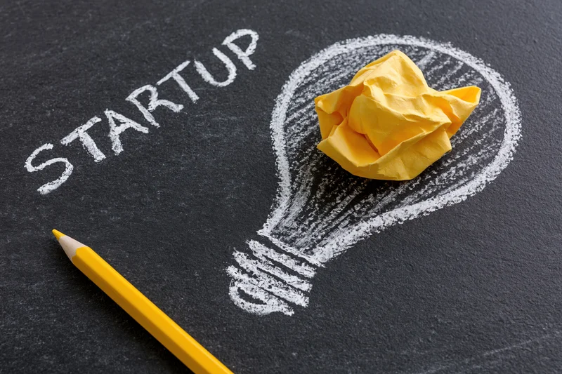 Chalk-drawn lightbulb with crumpled yellow paper inside and the word startup written beside it on a blackboard surface, with a yellow pencil nearby