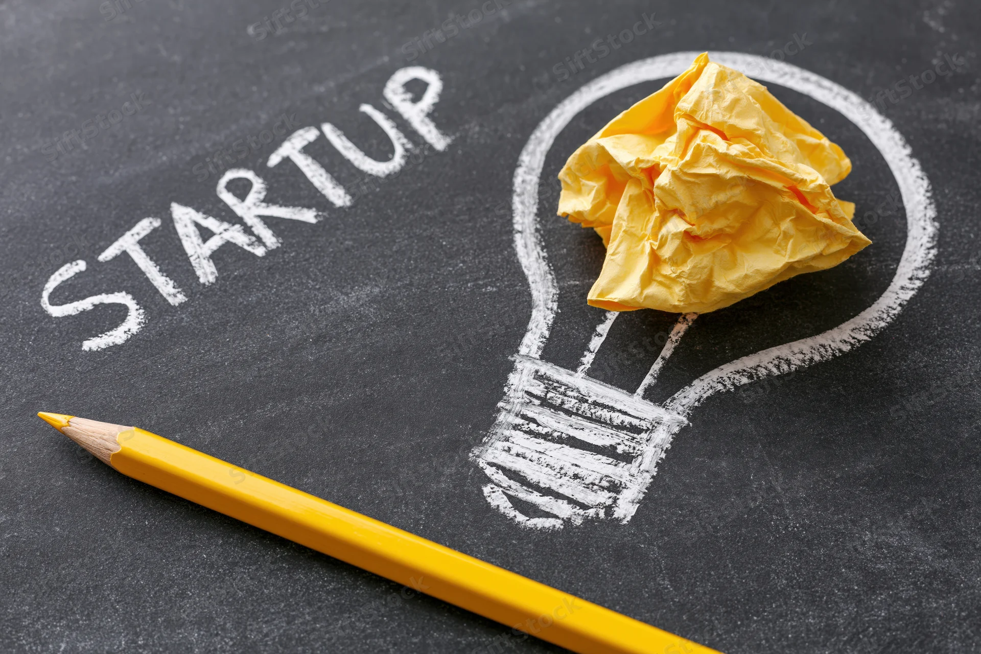 Chalk-drawn lightbulb with crumpled yellow paper inside and the word startup written beside it on a blackboard, with a yellow pencil in the foreground