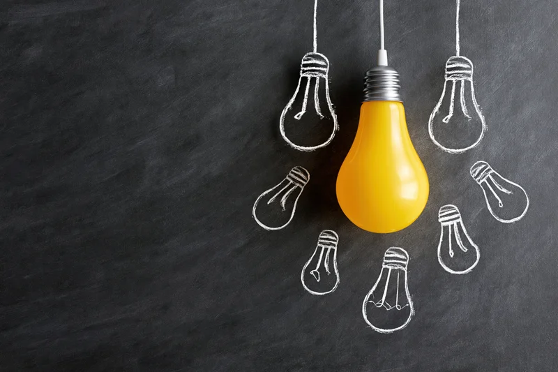 Yellow lightbulb hanging among chalk-drawn bulbs on a dark blackboard surface