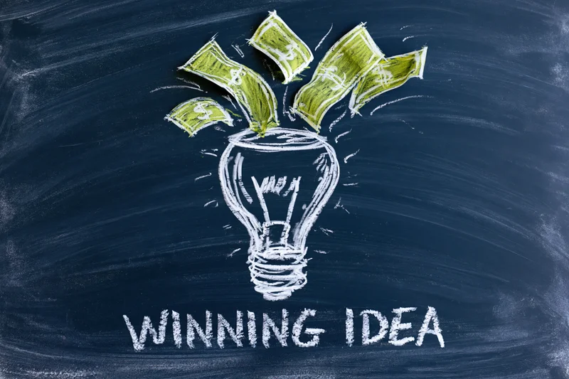 Chalk drawing of a lightbulb with money emerging from it on a dark navy blackboard with the words winning idea below