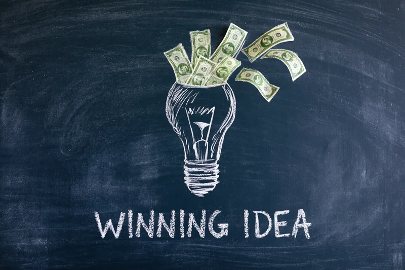 Chalk drawing of a lightbulb filled with dollar bills on a dark navy blackboard with the words winning idea written below