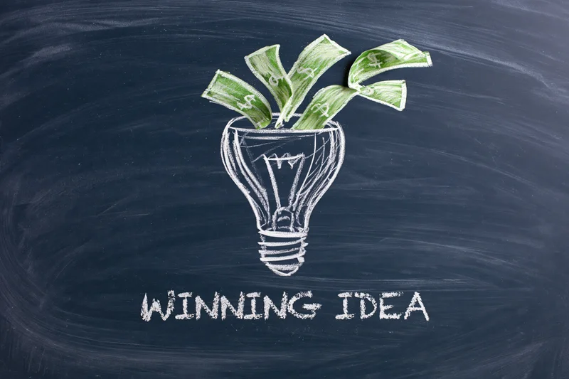 Chalk drawing of a lightbulb with money growing from it on a dark navy blackboard, labeled winning idea