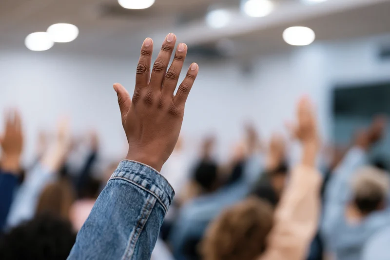 Raised hand in foreground with blurred group of students in classroom setting