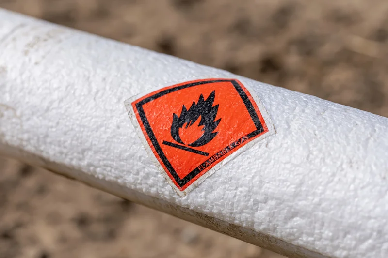 Close-up of orange diamond-shaped flammable gas hazard warning label on white textured pipeline outdoors