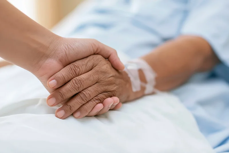 Younger hand holding elderly patient's hand in hospital bed