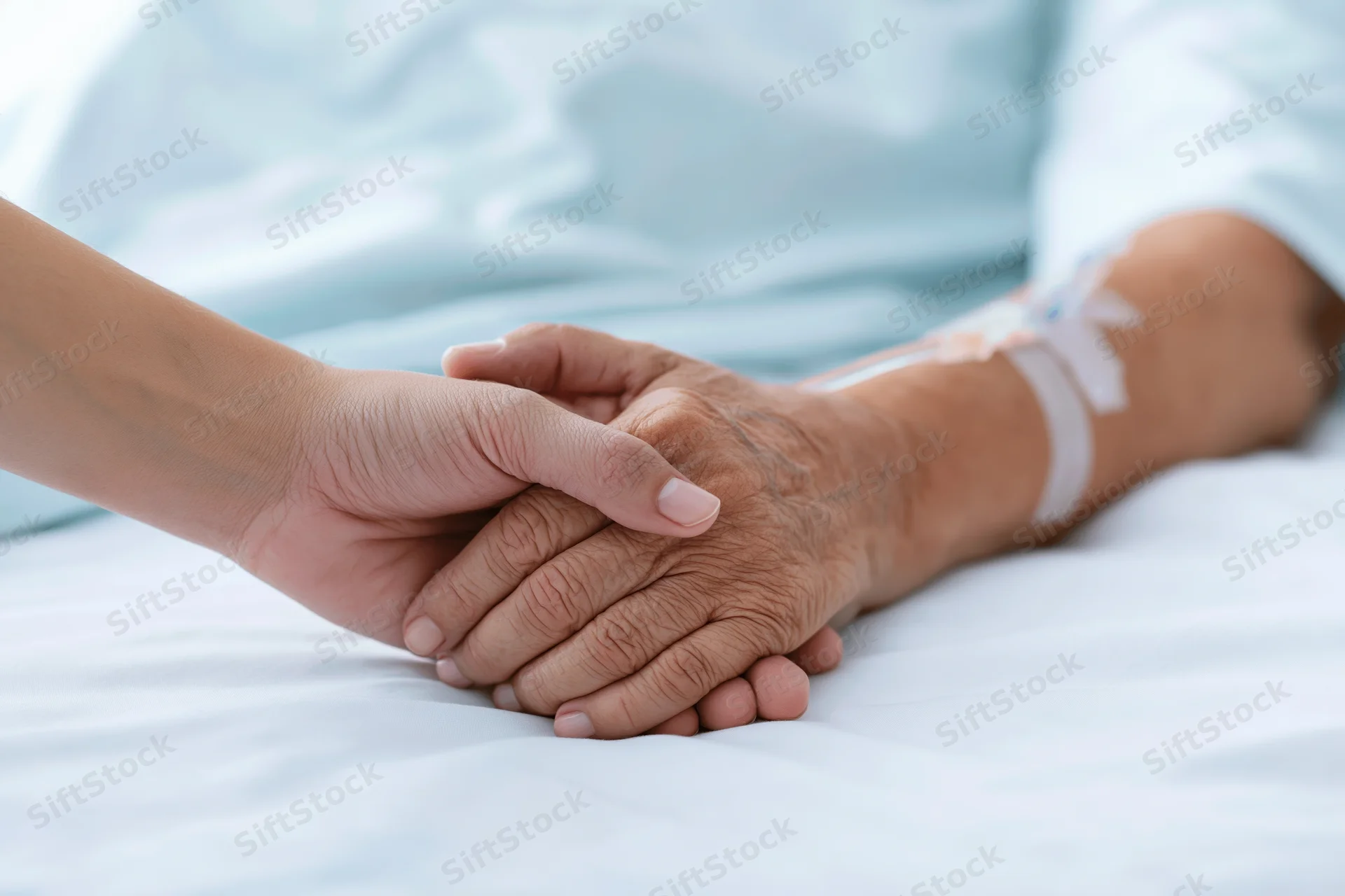 Younger hand gently holding elderly patient's hand on hospital bed