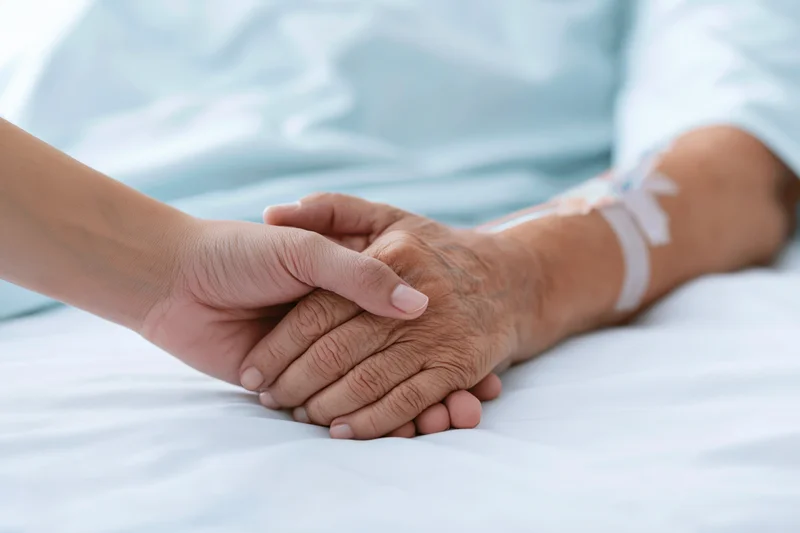 Younger hand gently holding elderly patient's hand on hospital bed