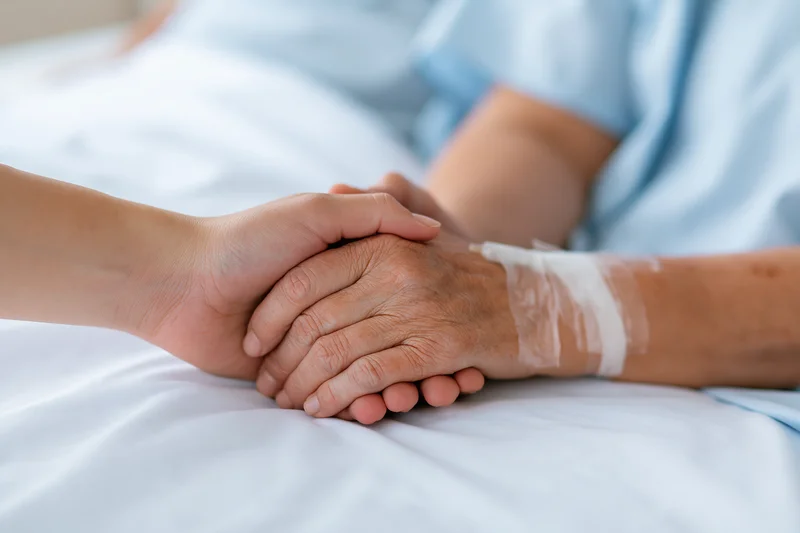 Younger hand holding elderly patient's hand on hospital bed