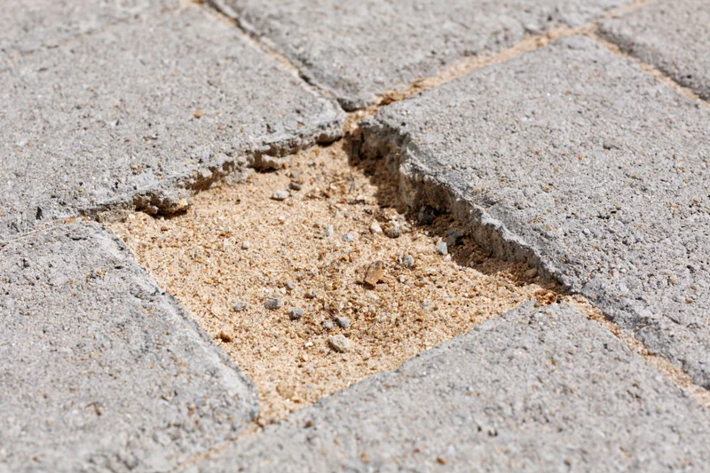 Damaged concrete brick pavement with missing paver and exposed sand in the gap
