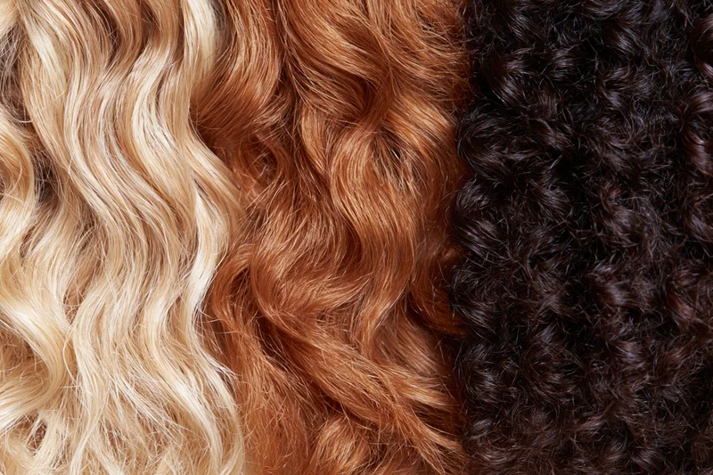 Close-up of three hair texture samples arranged side by side showing blonde wavy, brown wavy, and black curly hair