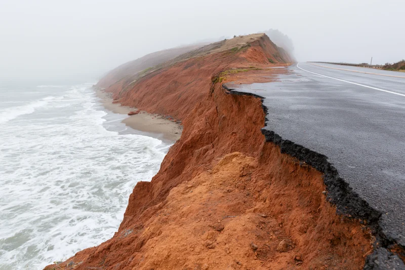Coastal road with eroded cliff edge next to ocean waves under foggy sky