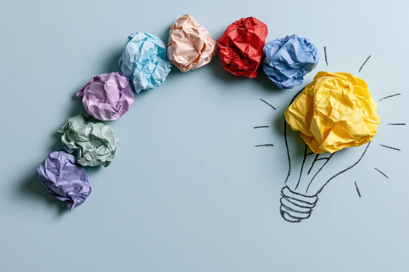 Colorful crumpled paper balls arranged in arc leading to hand-drawn lightbulb on light blue surface