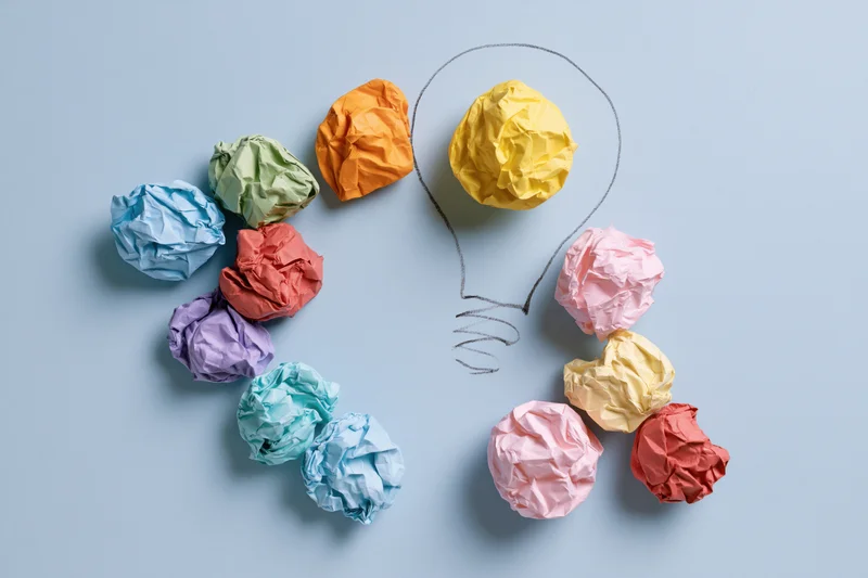 Colorful crumpled paper balls arranged around a hand-drawn lightbulb on a light blue background