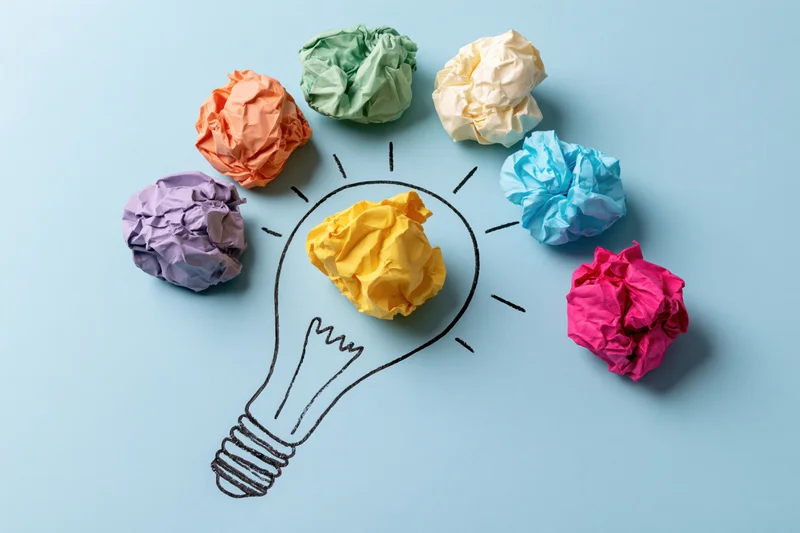 Crumpled colorful paper balls arranged around a hand-drawn lightbulb on a light blue surface