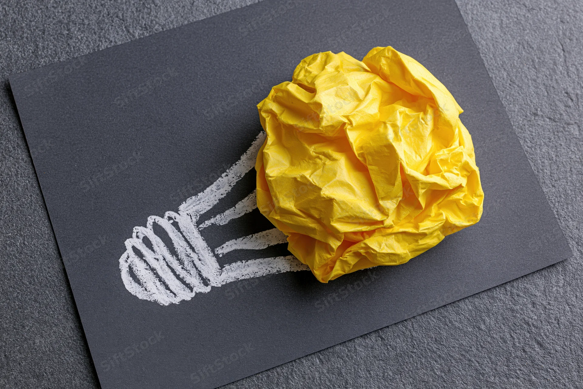 Crumpled yellow paper ball placed as bulb of hand-drawn lightbulb on dark board