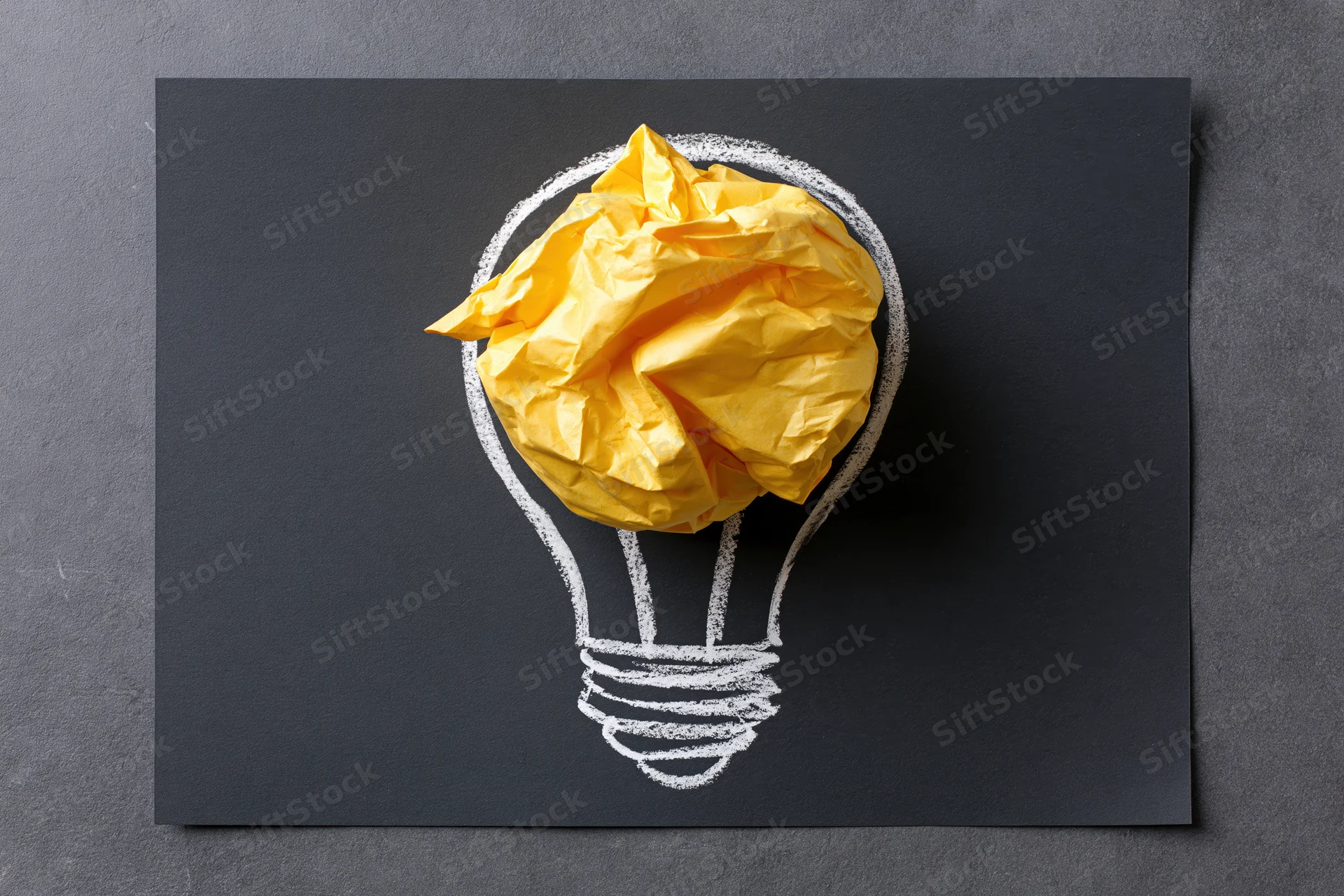 Crumpled yellow paper ball placed inside a chalk-drawn lightbulb on a black surface