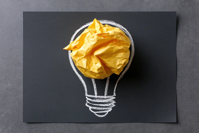 Crumpled yellow paper ball placed inside a chalk-drawn lightbulb on a black surface
