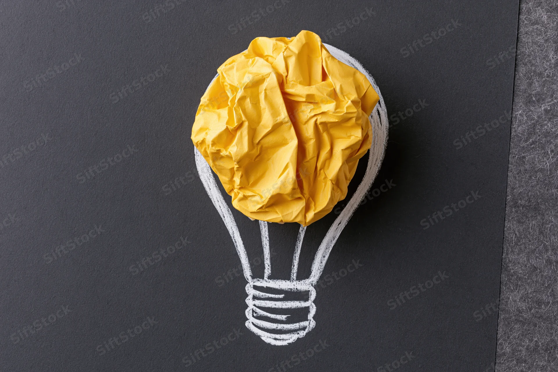 Crumpled yellow paper ball placed inside a chalk-drawn lightbulb on a black surface