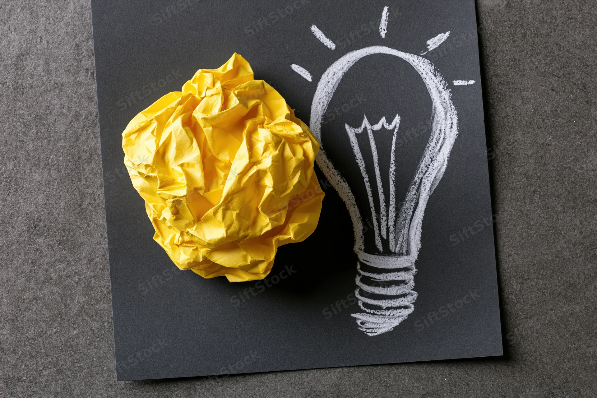 Crumpled yellow paper ball placed beside a chalk-drawn lightbulb on a black surface