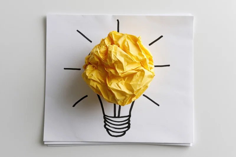 Crumpled yellow paper ball forming a lightbulb on a hand-drawn sketch on white paper