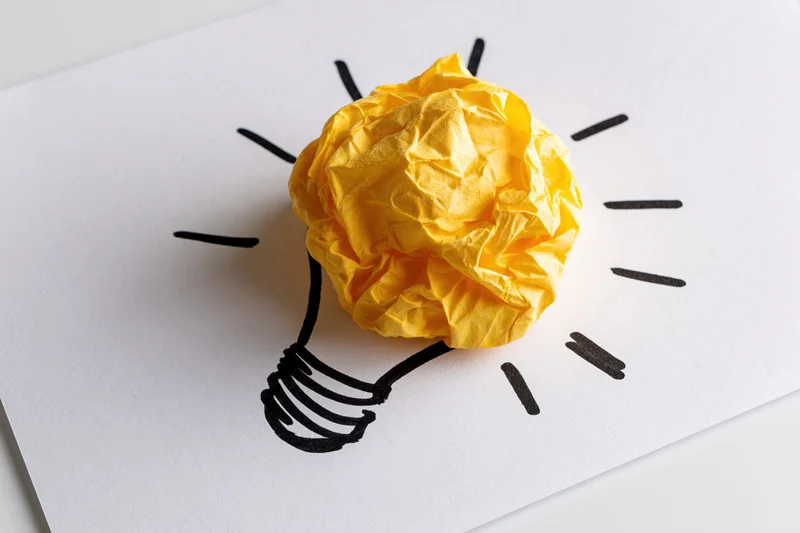Crumpled yellow paper ball forming a lightbulb on a hand-drawn sketch on white paper