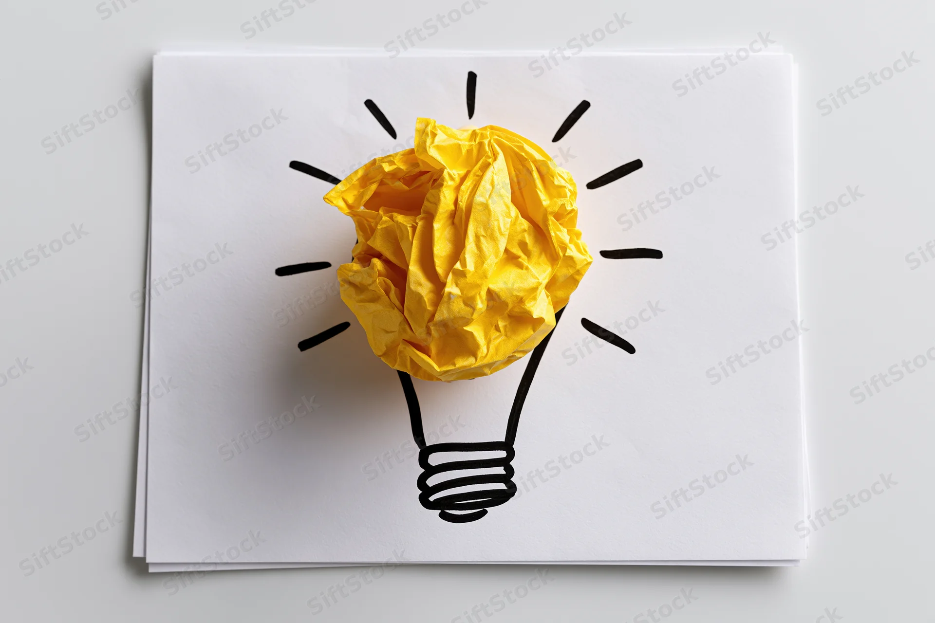 Crumpled yellow paper ball forming a lightbulb shape on a white sheet with hand-drawn lines