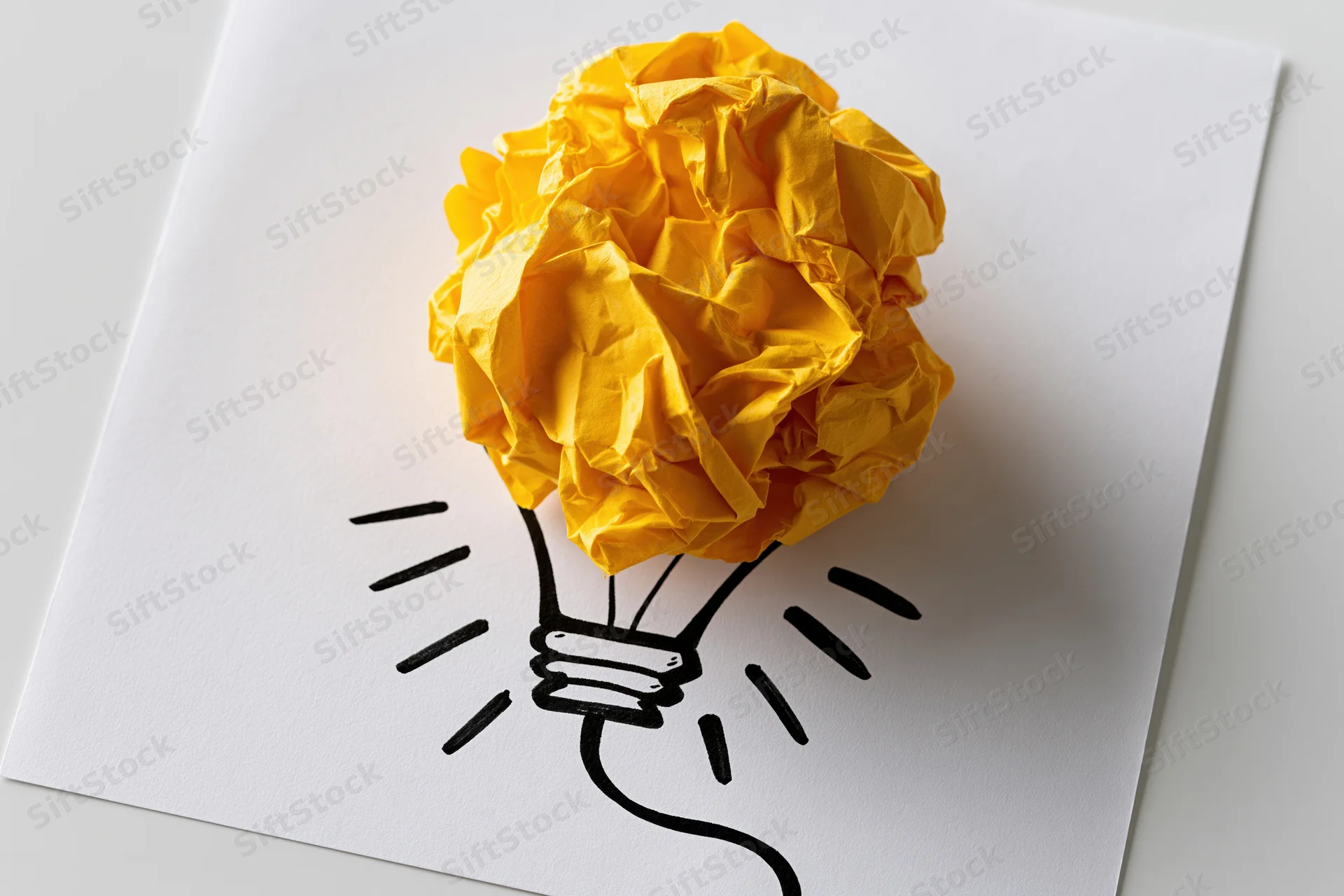 Crumpled yellow paper ball placed on a hand-drawn lightbulb sketch on white paper