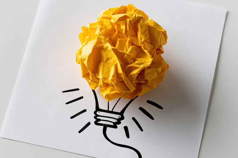 Crumpled yellow paper ball placed on a hand-drawn lightbulb sketch on white paper