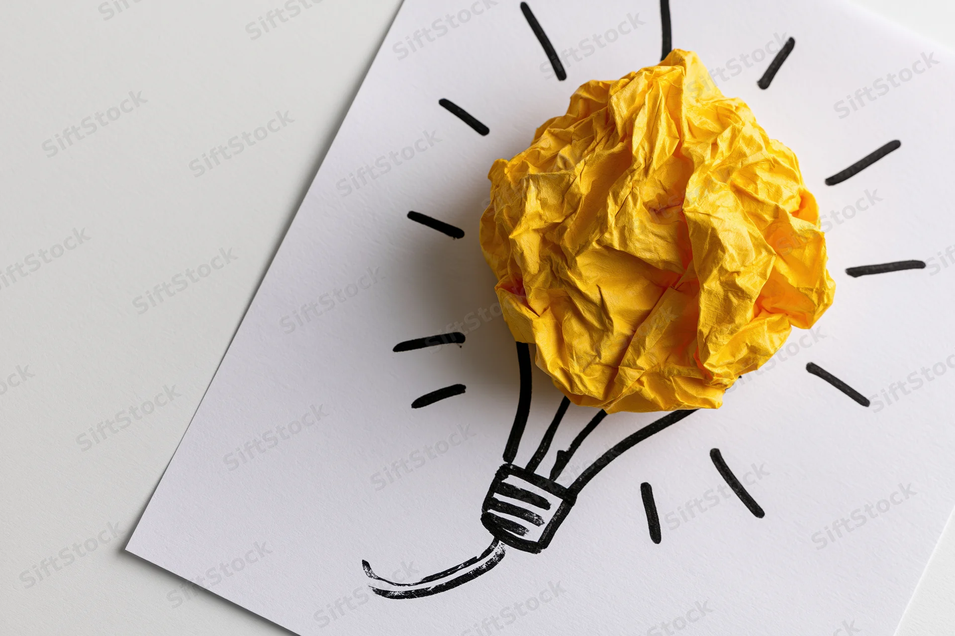 Crumpled yellow paper ball placed on a hand-drawn lightbulb sketch on white paper, symbolizing a new idea