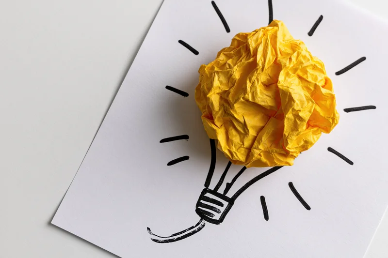 Crumpled yellow paper ball placed on a hand-drawn lightbulb sketch on white paper, symbolizing a new idea