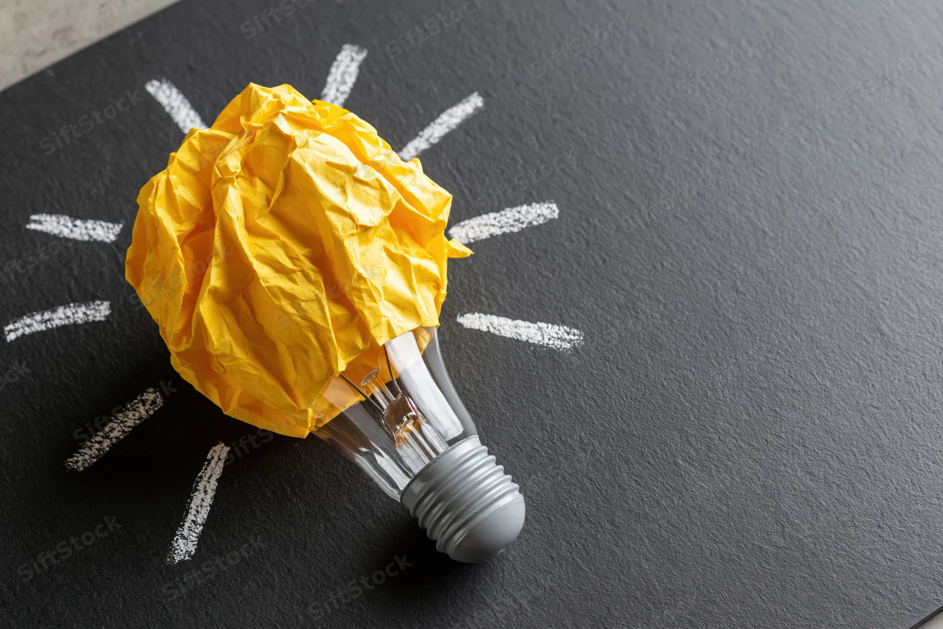 Crumpled yellow paper ball on a lightbulb with chalk rays on dark surface