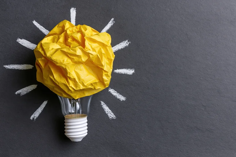 Crumpled yellow paper ball on a lightbulb with chalk rays on a blackboard