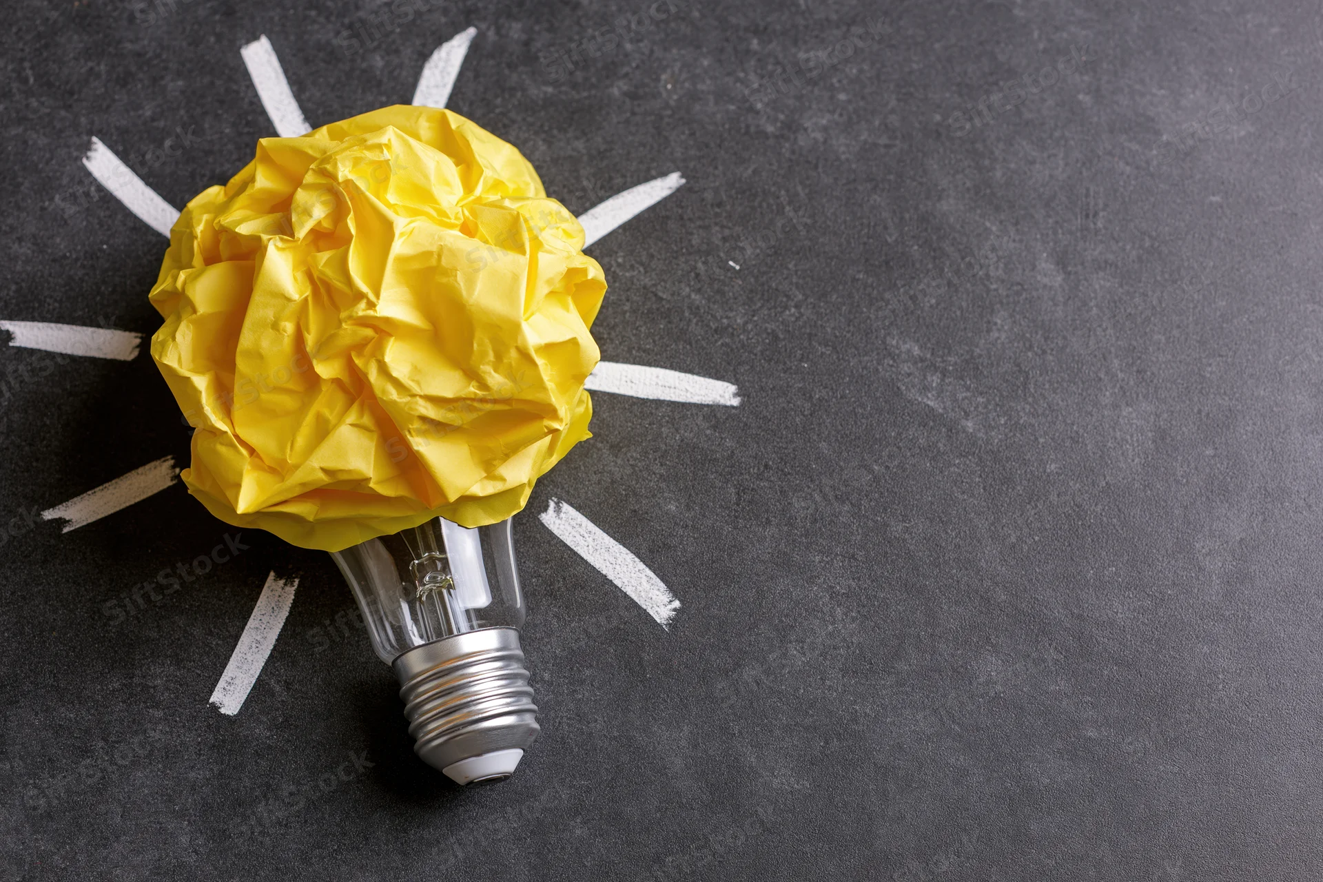 Crumpled yellow paper ball on a lightbulb with white rays drawn on dark surface