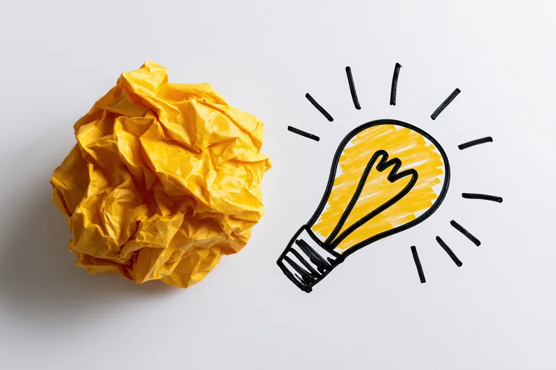 Crumpled yellow paper ball beside hand-drawn lightbulb on white surface