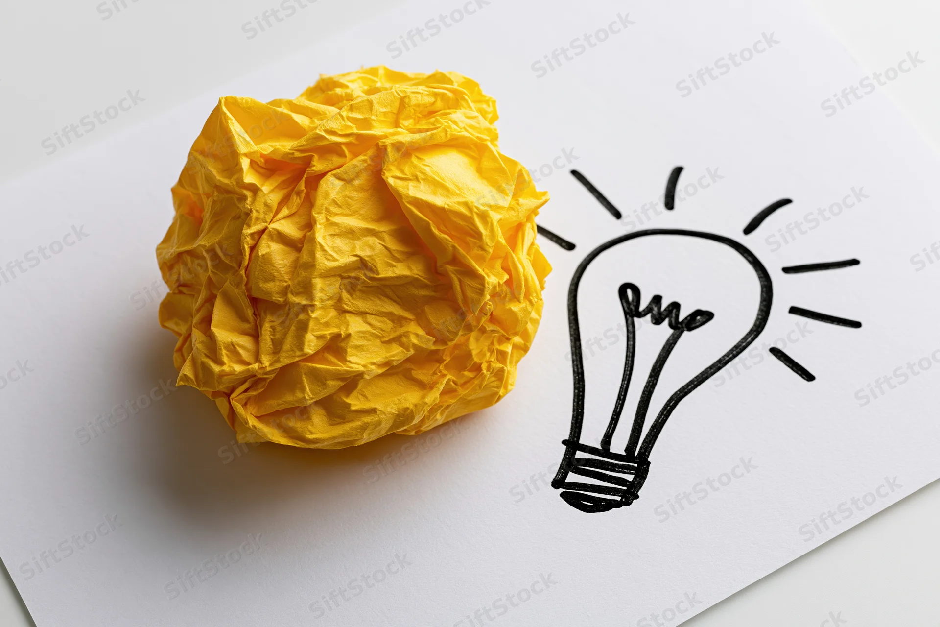 Crumpled yellow paper ball beside hand-drawn lightbulb on white sheet