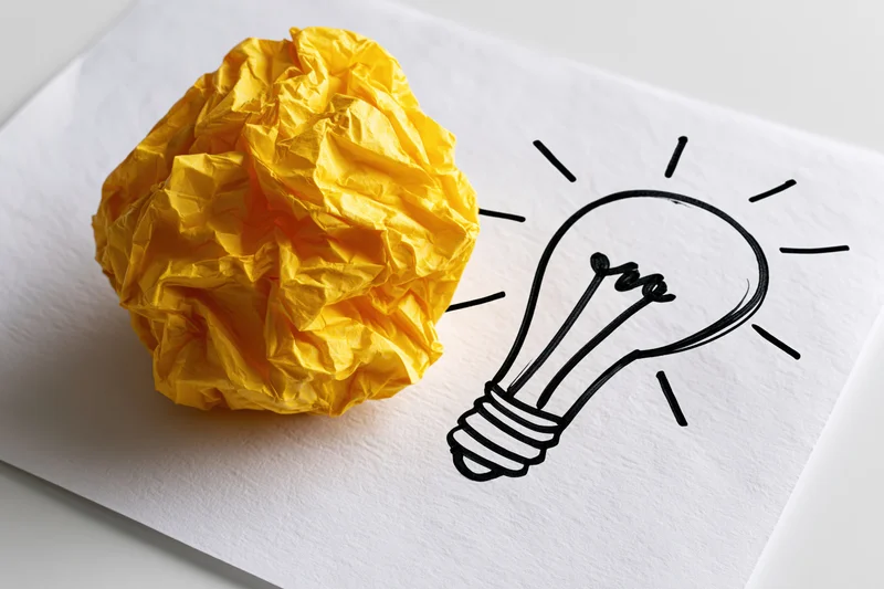 Crumpled yellow paper ball beside hand-drawn lightbulb on white sheet