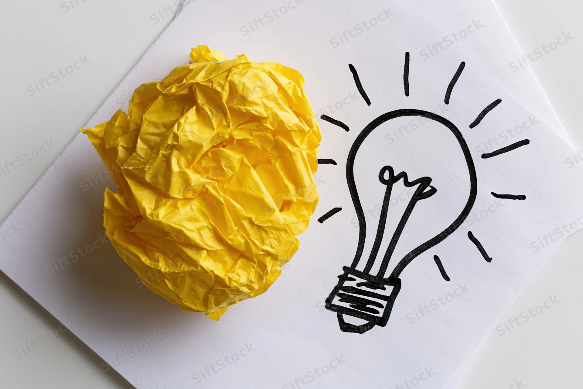 Crumpled yellow paper ball beside hand-drawn lightbulb on white paper sheet