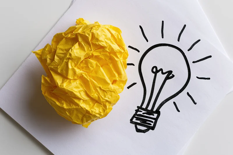 Crumpled yellow paper ball beside hand-drawn lightbulb on white paper sheet