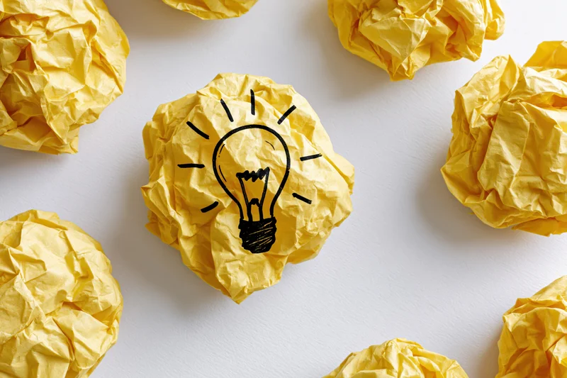 Crumpled yellow paper balls with a hand-drawn lightbulb on one, arranged on a white surface