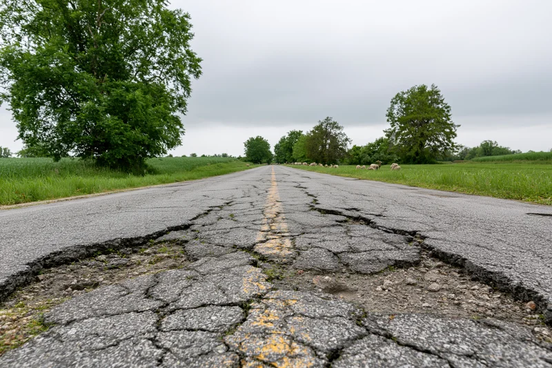 Damaged rural two-lane asphalt road stretching into the distance with cracked pavement and sheep grazing in adjacent fields